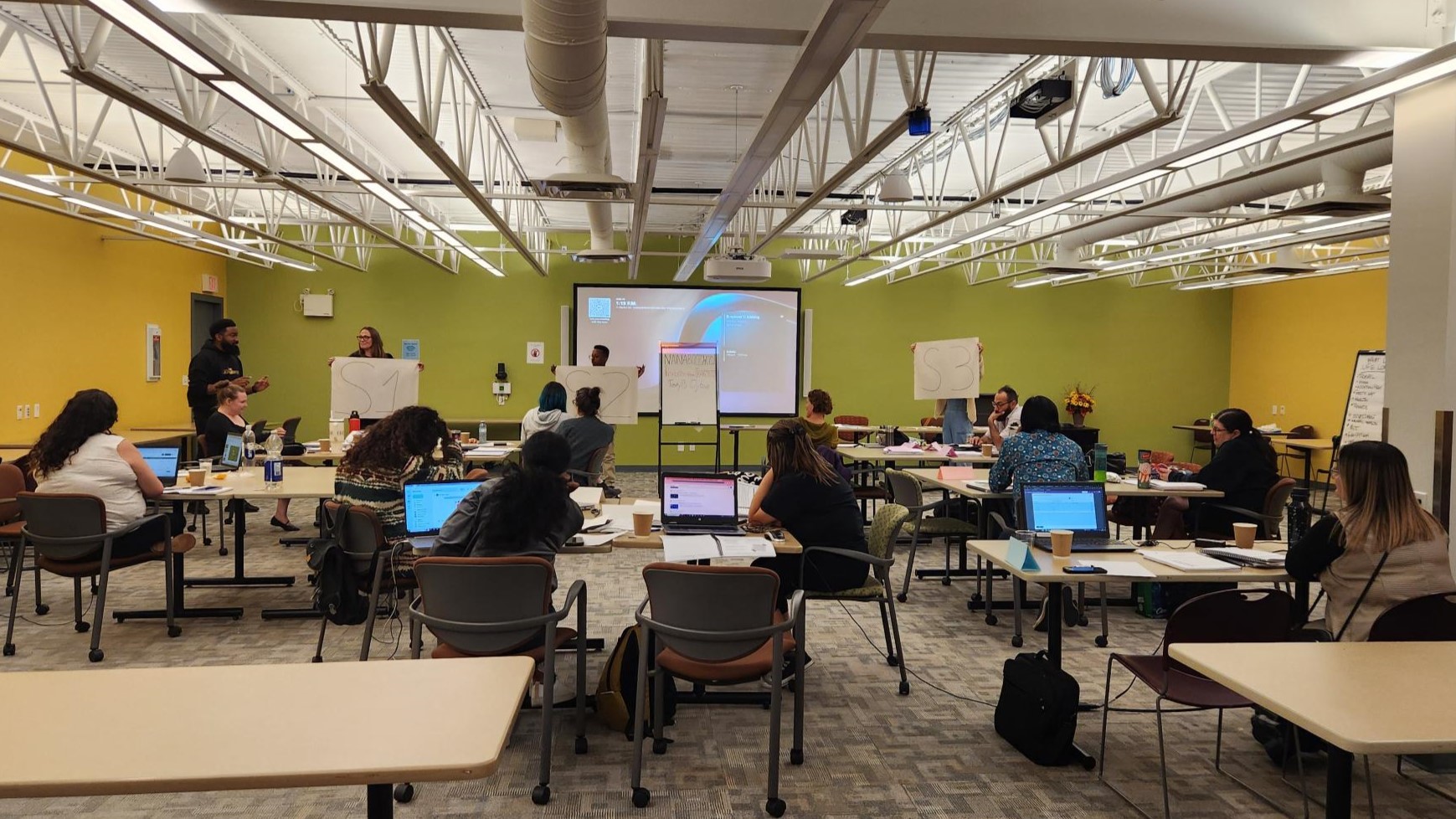 A group of students at desks in a classroom, photographed from behind, with four people presenting and holding flipchart paper at the front of the room.